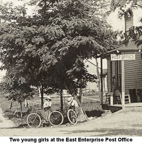 Black and white photograph showing two young girls in long skirts and broad straw hats standing with their fat-tired bicycles to the left of the porch of a building, from whose roof hangs a wooden sign reading 'POST OFFICE'; caption reads: 'Two young girls at the East Enterprise Post Office'